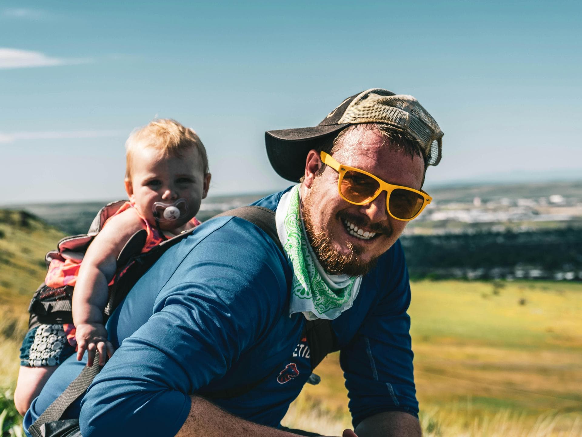 Auf einem Nordic-Walking-Familienausflug kann man sich mit dem Tragen des Babys abwechseln. Fröhlicher Vater trägt Baby auf dem Rücken durch die Landschaft.