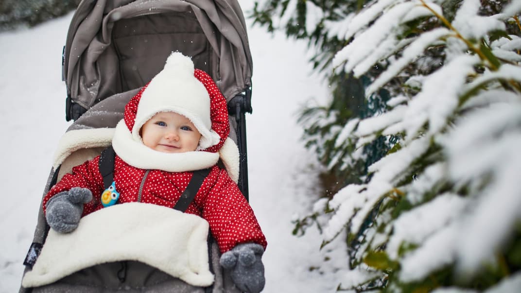 Ein kleines Mädchen sitzt dick eingepackt im Kinderwagen, der im Schnee steht.