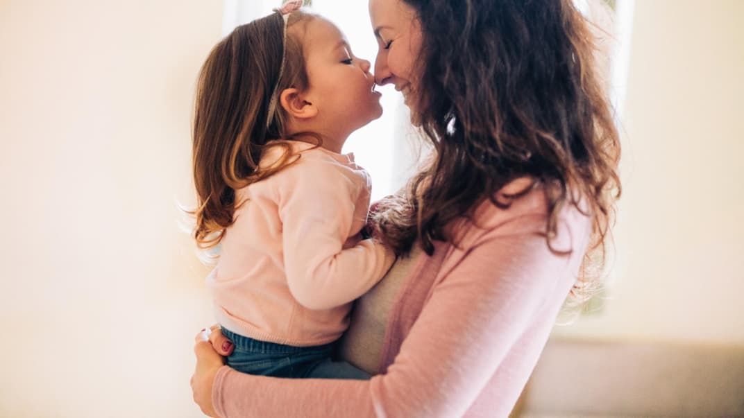 Eine Mutter trägt ihre kleine Tochter mit Schleife im Haar auf dem Arm.