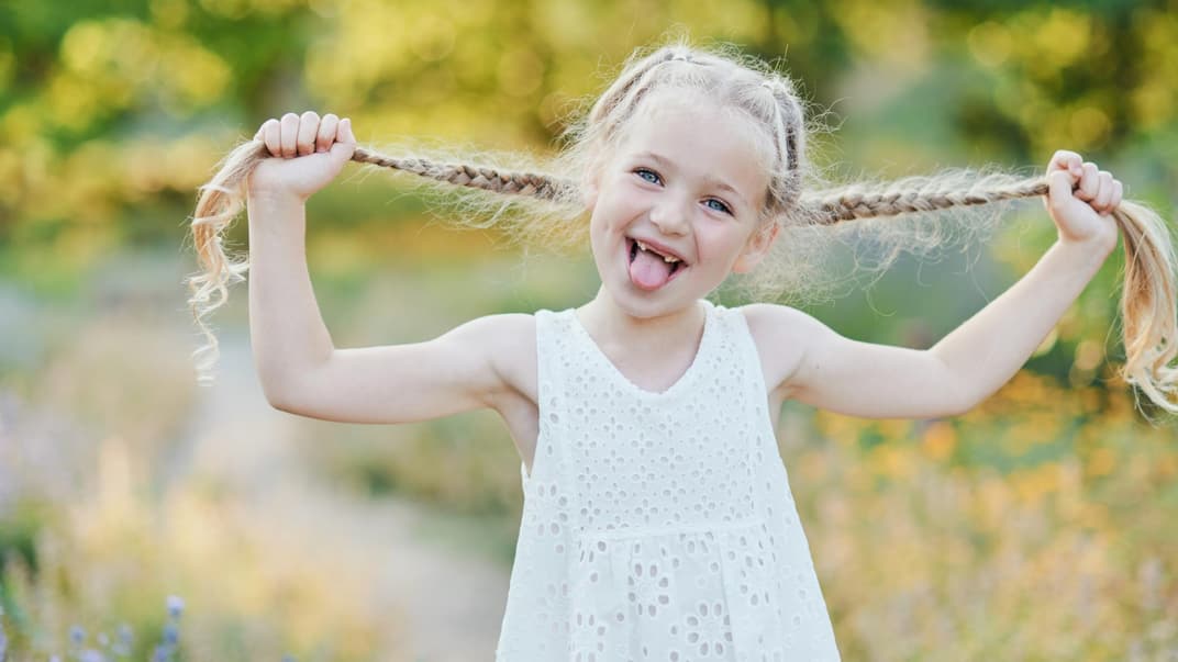 Ein kleines Mädchen in einem weißen Kleid, das seine Zöpfe zur Seite zieht.