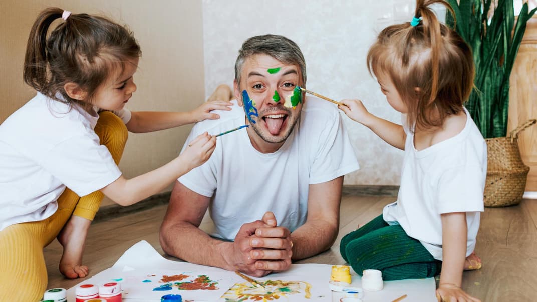 Ein männlicher Erzieher lässt sich in der Kita von zwei Kindern im Gesicht anmalen. 