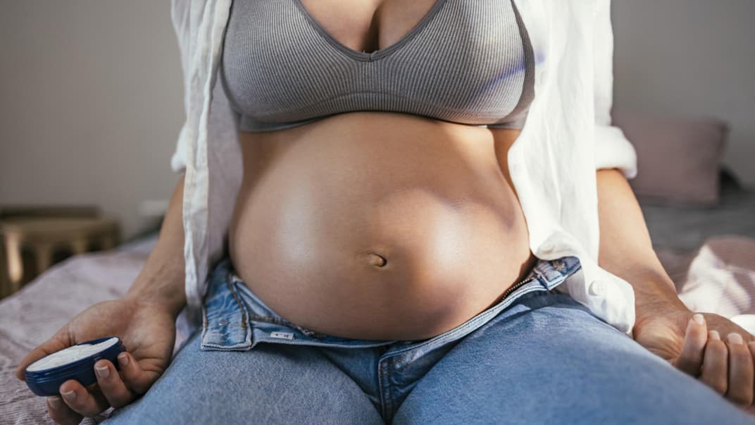 Eine Frau mit Babybauch sitzt mit geöffneter Hose auf dem Bett