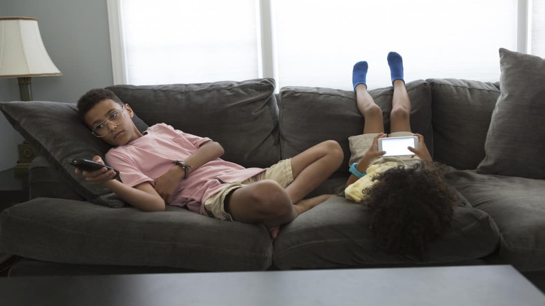Zwei Kinder liegen auf dem Sofa, eins mit Fernbedienung in der Hand, eins mit Smartphone