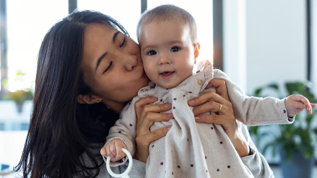 Eine asiatische Mutter hält ihr Baby-Mädchen hoch und küsst es zärtlich auf die Wange.