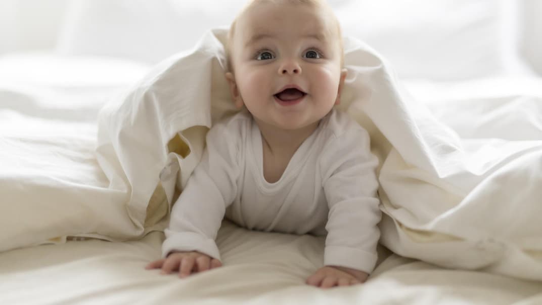 Ein kleiner Baby-Junge guckt frech unter einer weißen Bettdecke hervor. 