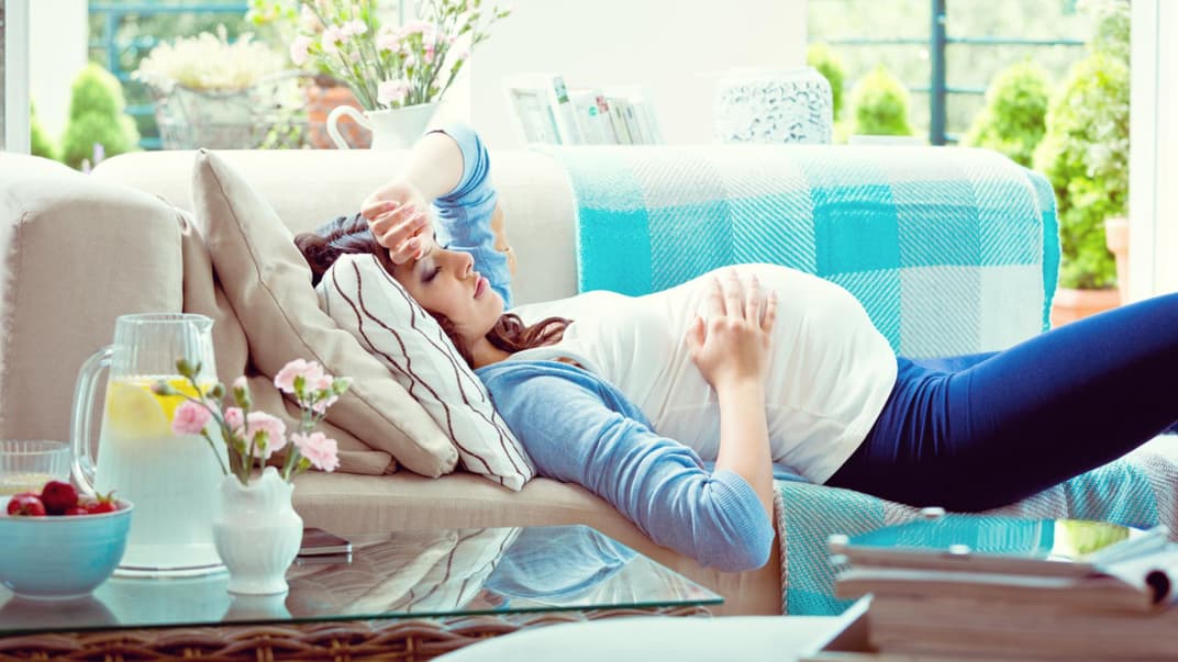 Schwangere Frau liegt auf dem Sofa und ruht sich aus – sie hat starke Schwangerschaftsbeschwerden.