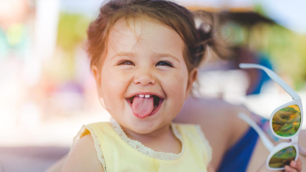 Ein kleines Mädchen mit Sonnenbrille in der Hand lacht und streckt die Zunge heraus.