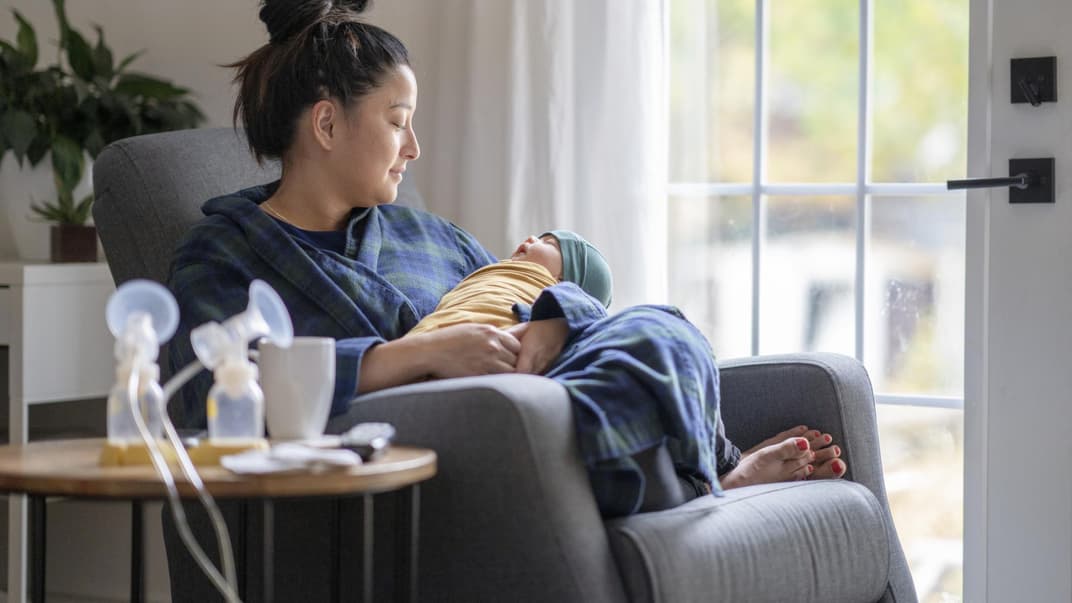Eine Mama sitzt mit ihrem Baby und einer Milchpumpe auf dem Sofa.