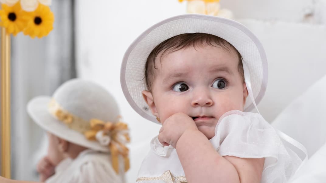 Ein kleines Baby-Mädchen mit Taufkleid und Hut schaut niedlich zur Seite. 