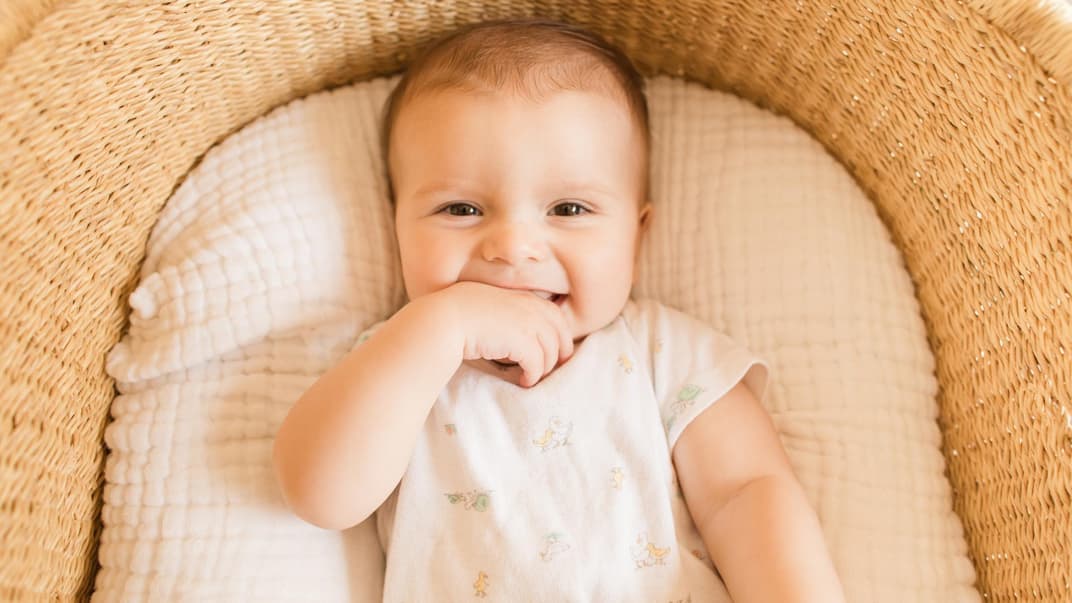 Ein kleiner, süßer Junge, der seine Finger im Mund hat und vom Babybett in die Kamera grinst.