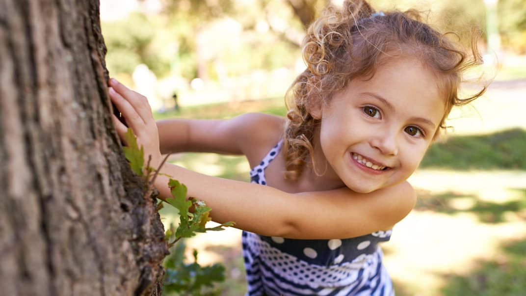 Ein kleines Mädchen schaut süß neben einem Baumstamm um die Ecke.