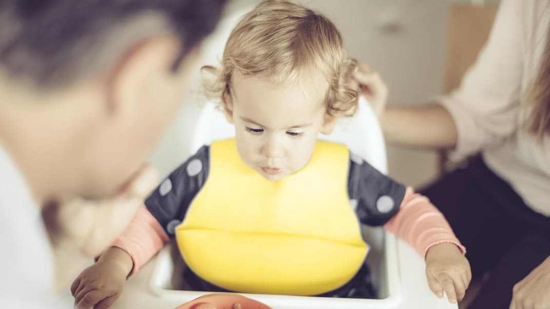Baby-Mädchen bekommt ihr Essen im Hochstuhl – das Lätzchen verhindert großes Chaos