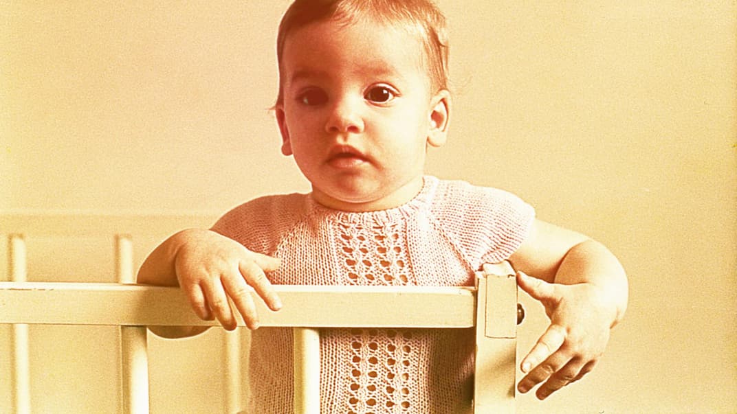 Ein Baby-Mädchen in den 60ern steht niedlich im Gitterbett