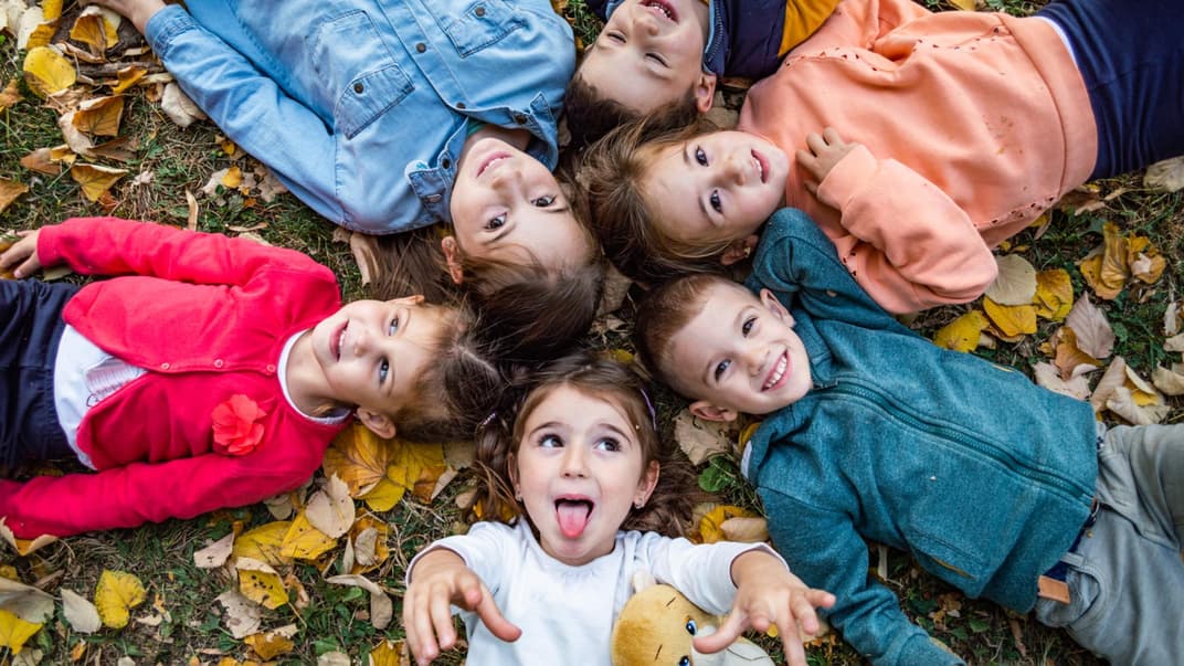 Kita-Kinder liegen im Herbstlaub und singen Herbstlieder im Kindergarten. 