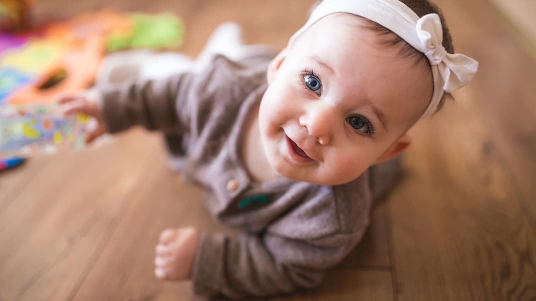 Kleines Baby-Mädchen mit Haarband liegt auf dem Boden und grinst.