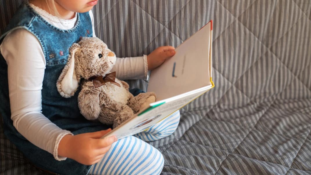 Kleines Mädchen sitzt auf einem Sofa, ein Hasen-Kuscheltier auf ihrem Schoß, und hält ein aufgeschlagenes Buch in der Hand.