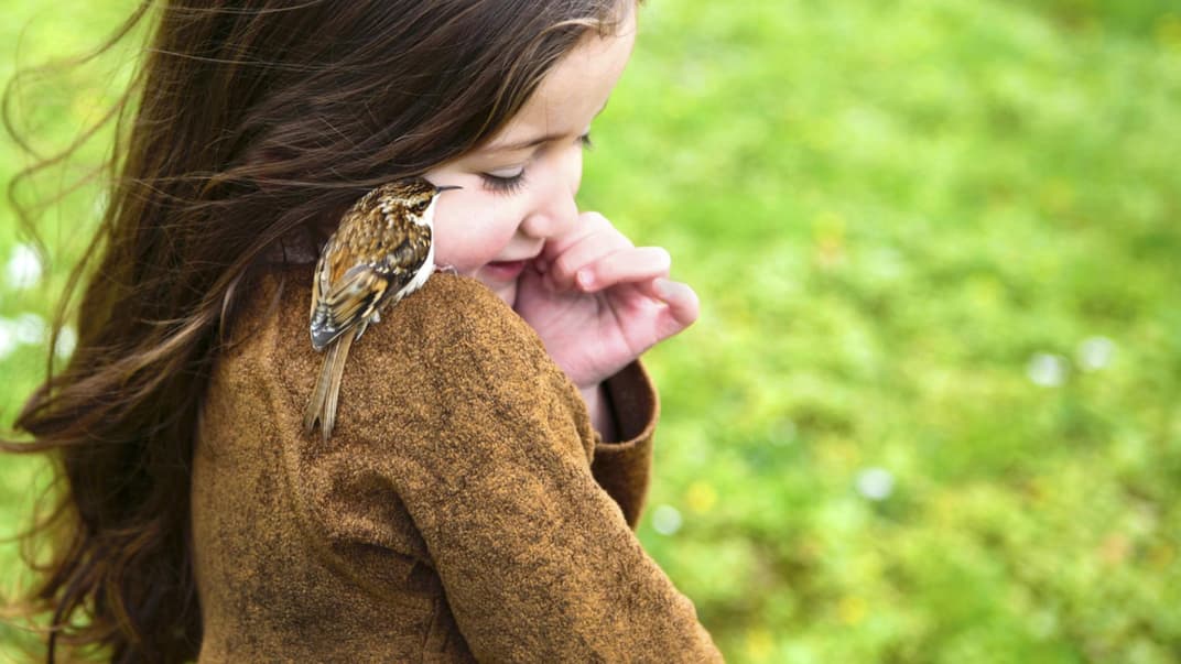 Ein Mädchen mit einem kleinen Vogel auf der Schulter steht auf einer Wiese.