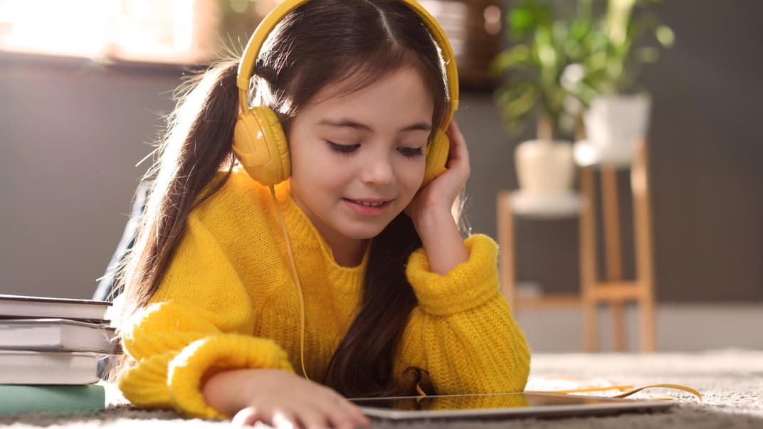 Ein Mädchen liegt auf dem Bauch auf dem Boden und hört ein Hörbuch mit Kopfhörern.