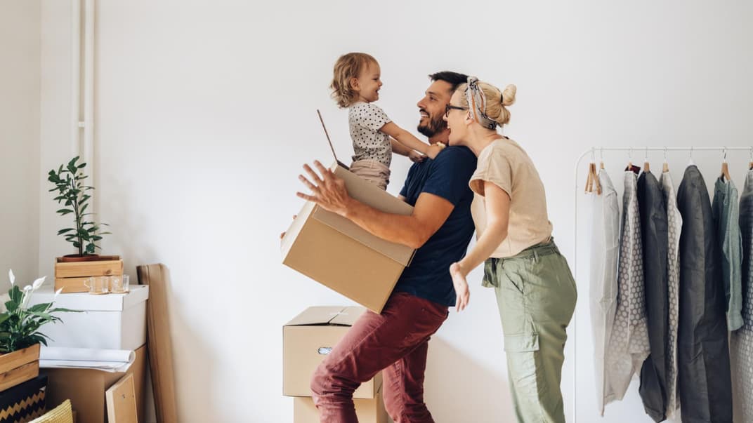 Fröhliche Familie zieht in ein neues Zuhause und das Kleinkind sitzt im Umzugskarton. 