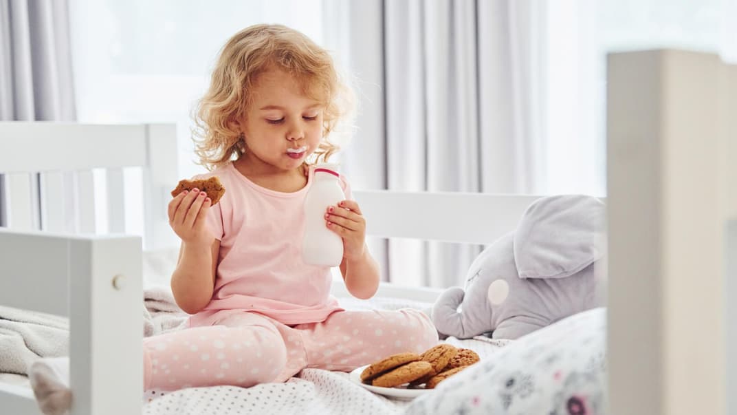Ein kleines Mädchen isst im Bett Kekse und trinkt Milch.