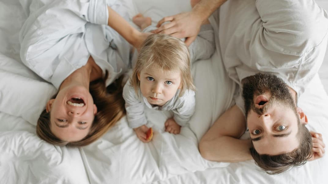 Eine glückliche Mutter liegt mit ihrem eher traurigen Kleinkind und neuen Partner im Bett.