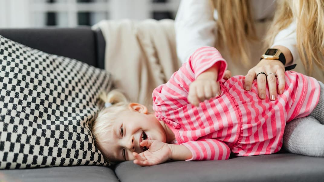 Ein kleines Mädchen wird von seiner Mutter auf dem Sofa gekitzelt.