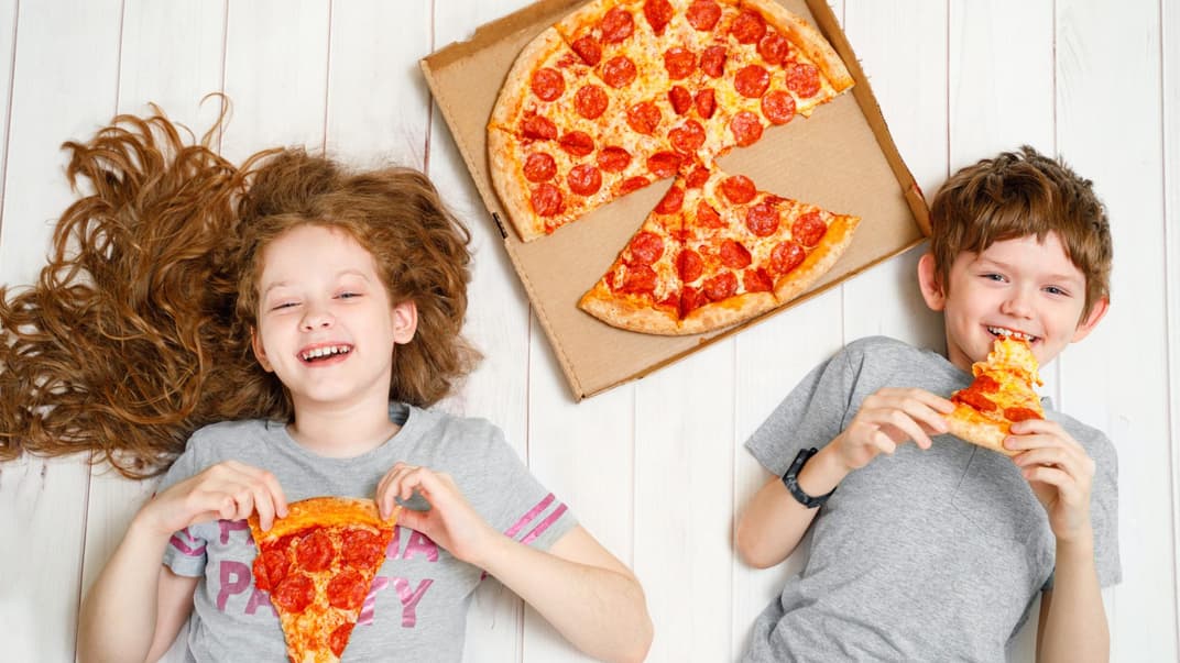 Zwei fröhliche Kinder liegen mit Salami-Pizza auf dem Boden.