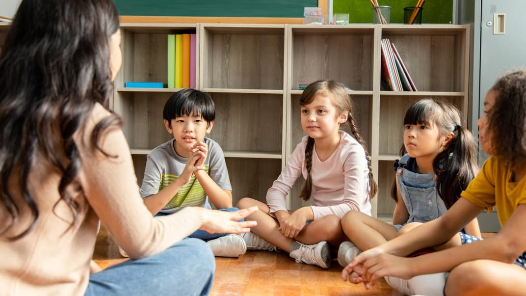 Lehrerin sitzt mit Grundschulkindern auf dem Boden.