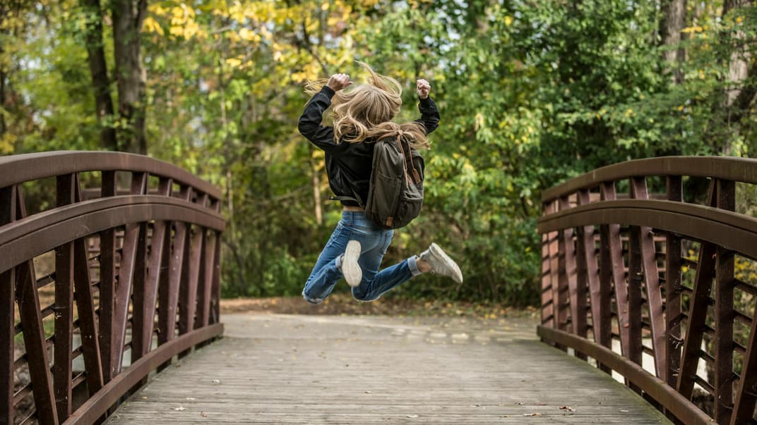 Mädchen mit Schulranzen springt hoch auf einer Holzbrücke.