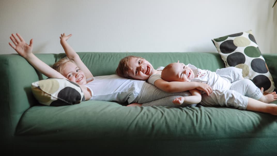 Drei lachende Geschwister liegen auf dem Sofa.