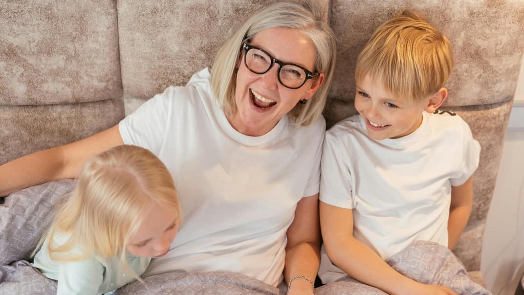 Eine Oma sitzt lachend mit ihren zwei Enkelkindern auf dem Sofa.