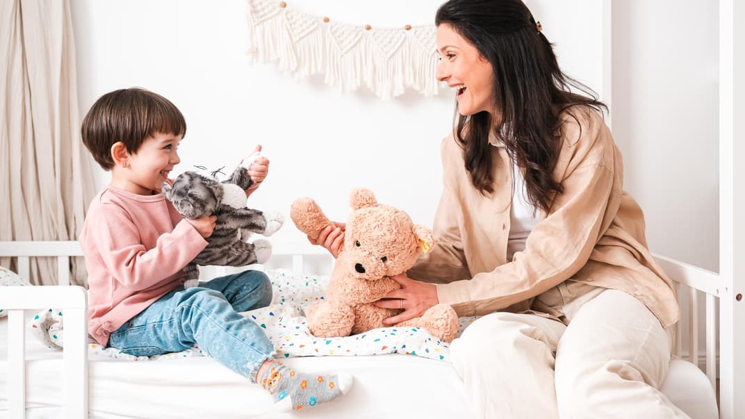 Mutter und Kind spielen auf Bett mit Kuscheltieren.