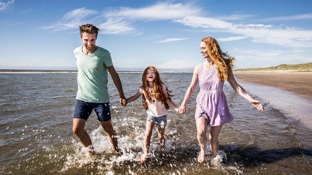 Ein Mädchen läuft mit ihren Eltern durch seichtes Meerwasser. 