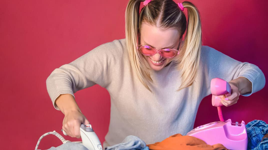 Fröhliche Frau mit pinker Brille bügelt und telefoniert gleichzeitig.