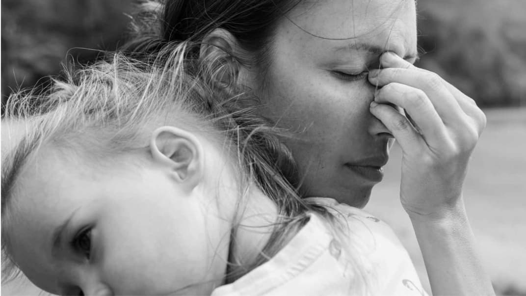 Eine verzweifelte Mutter hält ihre Tochter auf dem Arm, während sie die Stirn runzelt, die Augen schließt und sich an die Nase fässt.