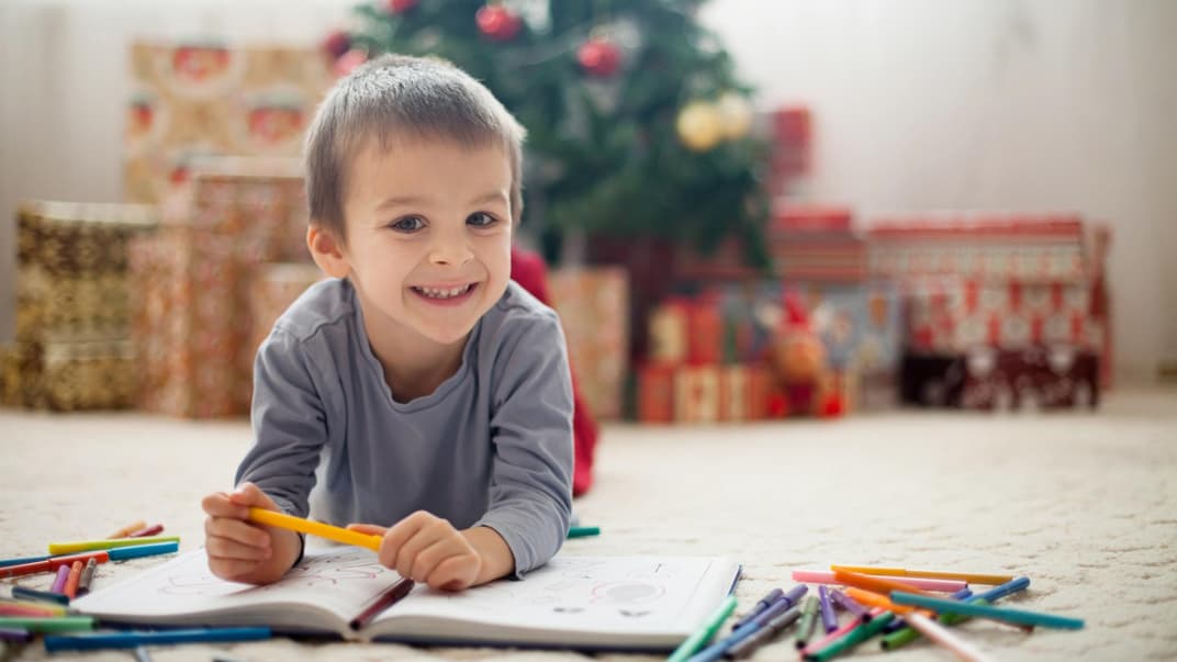 Ein kleiner Junge liegt vor dem Weihnachtsbaum auf dem Boden und malt Bilder..