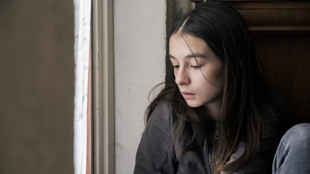 Ein junges Mädchen sitzt in ihrem Zimmer und schaut traurig aus dem Fenster.