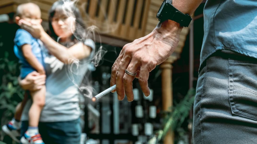 Eine Oma raucht vor einem Baby. Die Mutter hält ihm den Mund zu. 