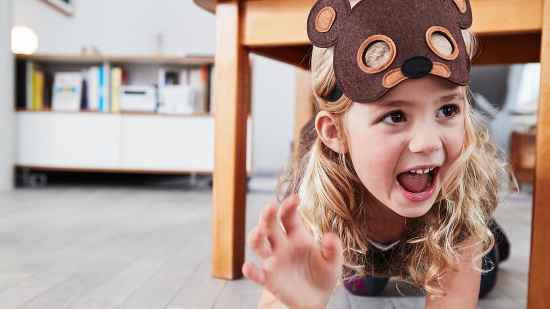 Ein kleines Mädchen spielt mit Bären-Maske unter dem Tisch.