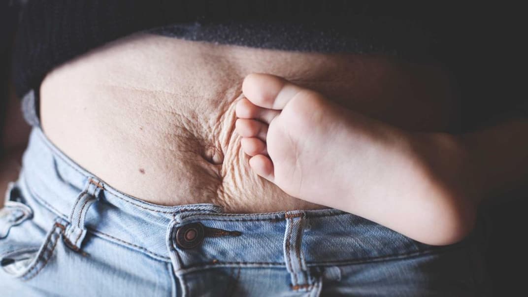 Ja, genau so darf ein After-Baby-Bauch aussehen. Kinderfuß an faltigem Mama-Bauch.