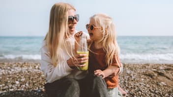 Mutter und Tochter sitzen fröhlich am schwedischen Strand, trinken einen Smoothie und lachen. 