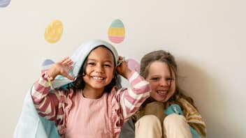 Zwei lachende Mädchen sitzen vor einer Wand, die mit Ostereierbildern beklebt ist. 