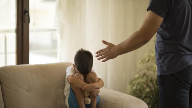 Junge sitzt mit angezogenen Knien auf dem Sofa, während sein Vater mit ihm schimpft.