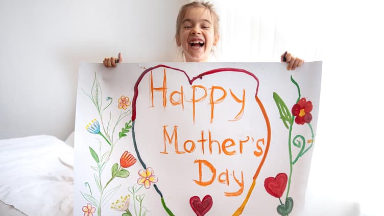 Ein kleines Mädchen hält ein selbst gebasteltes Bild mit der Aufschrift "Happy Mothers Day".