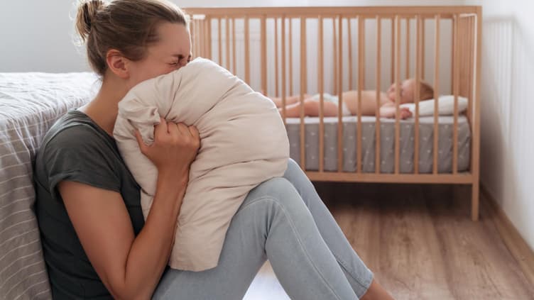 Eine Frau schreit in ein Kissen, während ihr Baby im Bett schläft