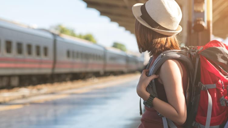 Ein junges Mädchen steht mit einem Rucksack an einem Bahngleis und wartet.
