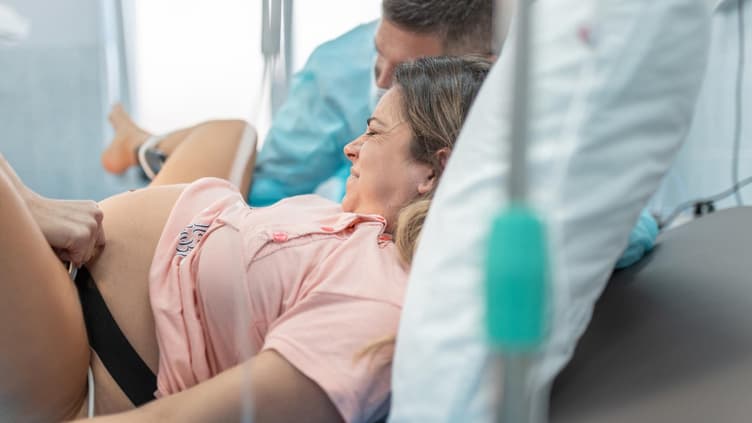 Frau liegt im Bett unter der Geburt, Mann sitzt neben ihr.