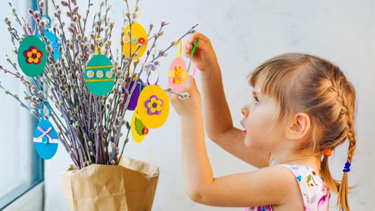 Kleines Mädchen hängt selbst gebastelte Ostereier an den Strauch.