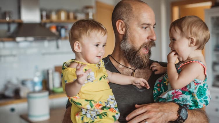 Papa mit Vollbart hat zwei Kinder auf dem Arm.