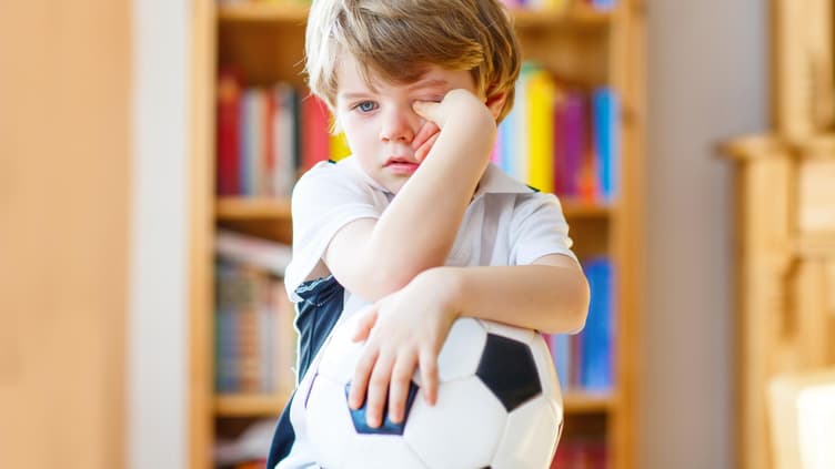 Krankes Kind reibt sich die Augen mit Fußball im Arm.